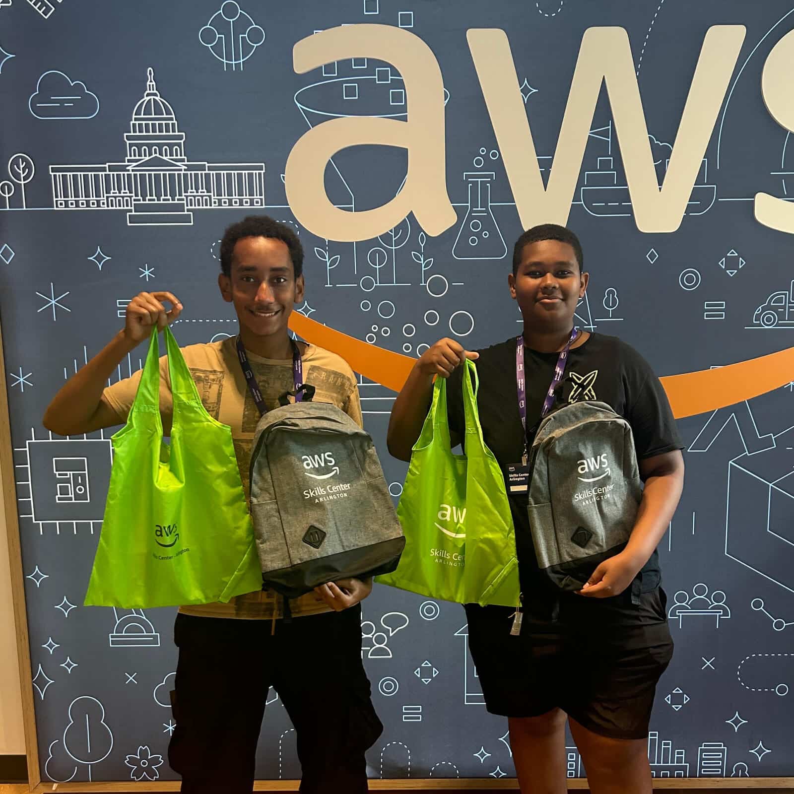 Two smiling young people stand in front of an AWS backdrop, each holding a green tote bag and a gray AWS backpack featuring the Affordable Homes & Communities (AHC) logo, looking happy with their event swag.