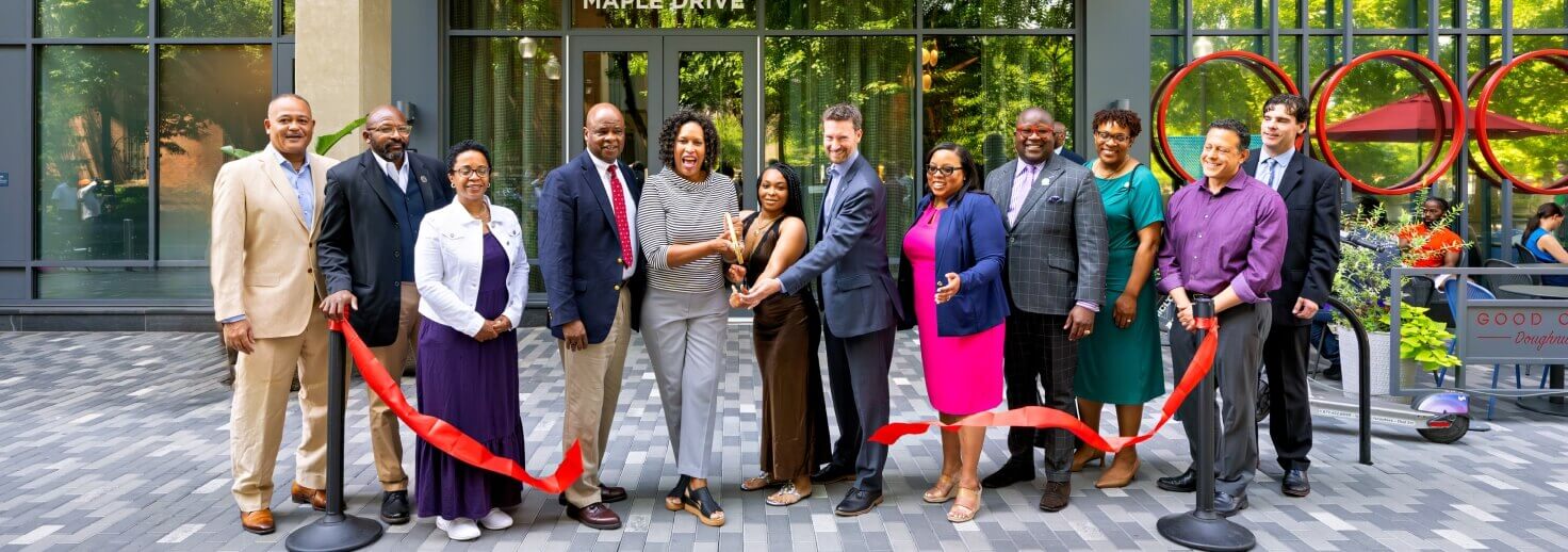 A group of people in business and formal attire stand smiling in front of a building, holding a red ribbon for an Affordable Homes & Communities Leadership ribbon-cutting ceremony. Two people in the center are cutting the ribbon.