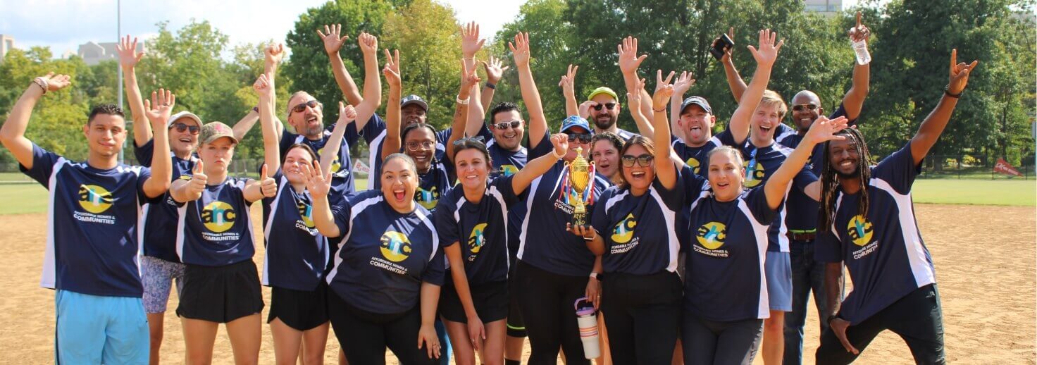 A group of people in matching blue sports shirts pose outdoors on a baseball field, smiling and raising their arms in celebration. Showcasing Affordable Homes & Communities Leadership, some hold a trophy, with trees visible in the background.