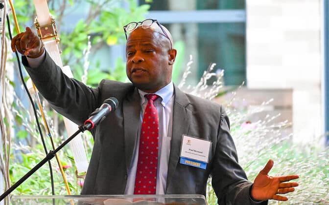 A man in a suit and red tie speaks at a podium with a microphone, gesturing with one hand raised and wearing glasses on his head. A name tag reading President is visible on his jacket.