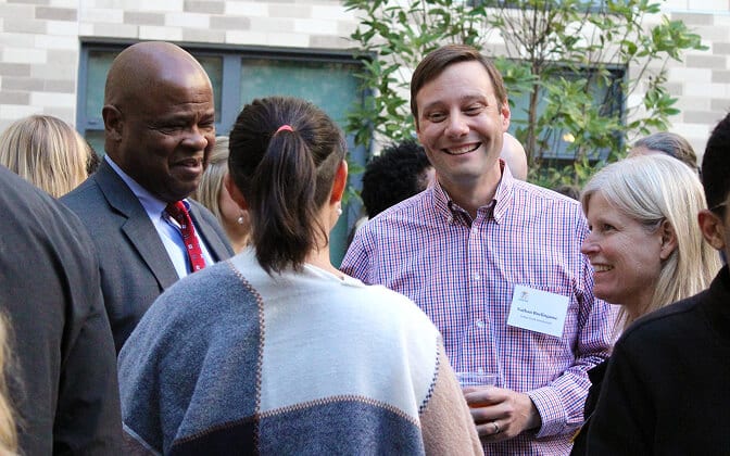 A group of people are talking and smiling at an outdoor event. One man is wearing a suit, while others are dressed casually. The background shows a building and some greenery.