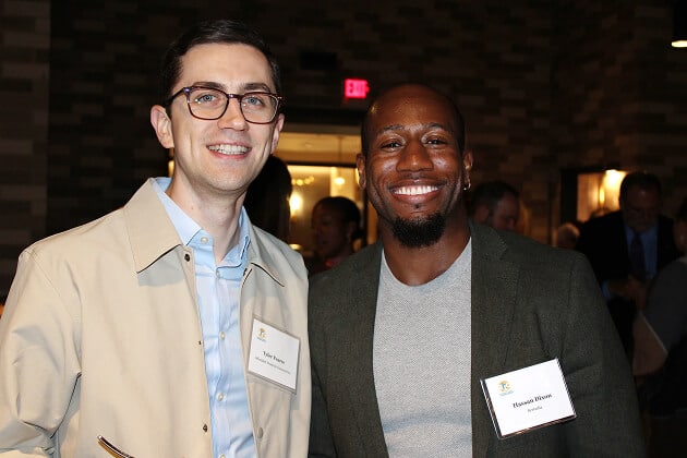 Two men wearing nametags smile for a photo at an indoor event. The man on the left wears glasses, a light jacket, and a blue shirt. The man on the right wears a grey blazer and white shirt. People and lights are visible in the background.