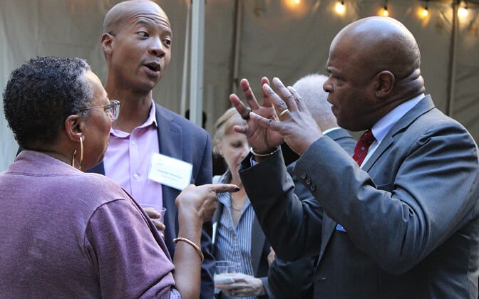 Three people in business attire are engaged in an animated conversation at an outdoor event; one man gestures expressively while the others listen intently. String lights are visible in the background.