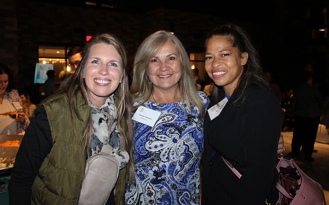 Three women smile and pose together at an indoor event. They wear nametags and casual to semi-formal clothing. Other people and tables with food are visible in the dimly lit background.