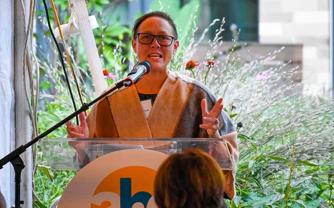 A person wearing glasses and a tan shawl speaks at a podium with a microphone, gesturing with their hands. The podium has a circular logo, and greenery and flowers are visible in the background.