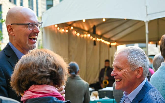 Two men in suits are smiling and laughing at an outdoor event, with string lights and a tent in the background. A woman with brown hair and a pink scarf stands with her back to the camera, engaged in the conversation.