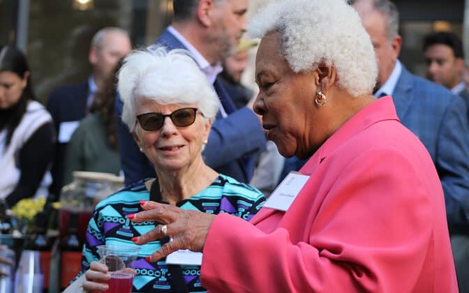 Two older women talk at an outdoor social event. One wears sunglasses and a blue patterned top, holding a drink, while the other wears a bright pink blazer and gestures with her hand. Other people are blurred in the background.
