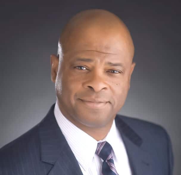 A man in a dark suit, light dress shirt, and striped tie poses for a professional headshot against a neutral gradient background. He is bald and slightly smiling.
