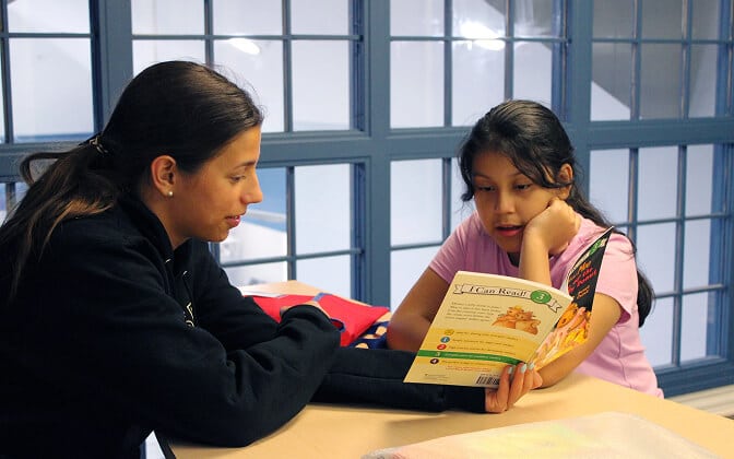 Two girls sit at a table by a window. One girl attentively listens while the other, wearing a pink shirt, reads aloud from a colorful children’s book.