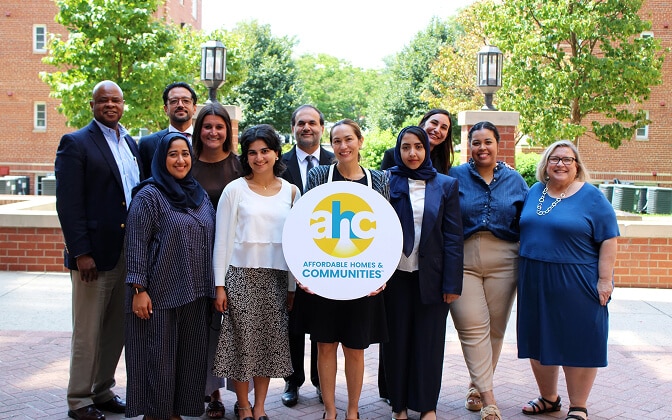Thirteen people smile and stand outdoors in business and casual attire, holding a round sign that reads AHC Affordable Homes & Communities,” with buildings and trees in the background.