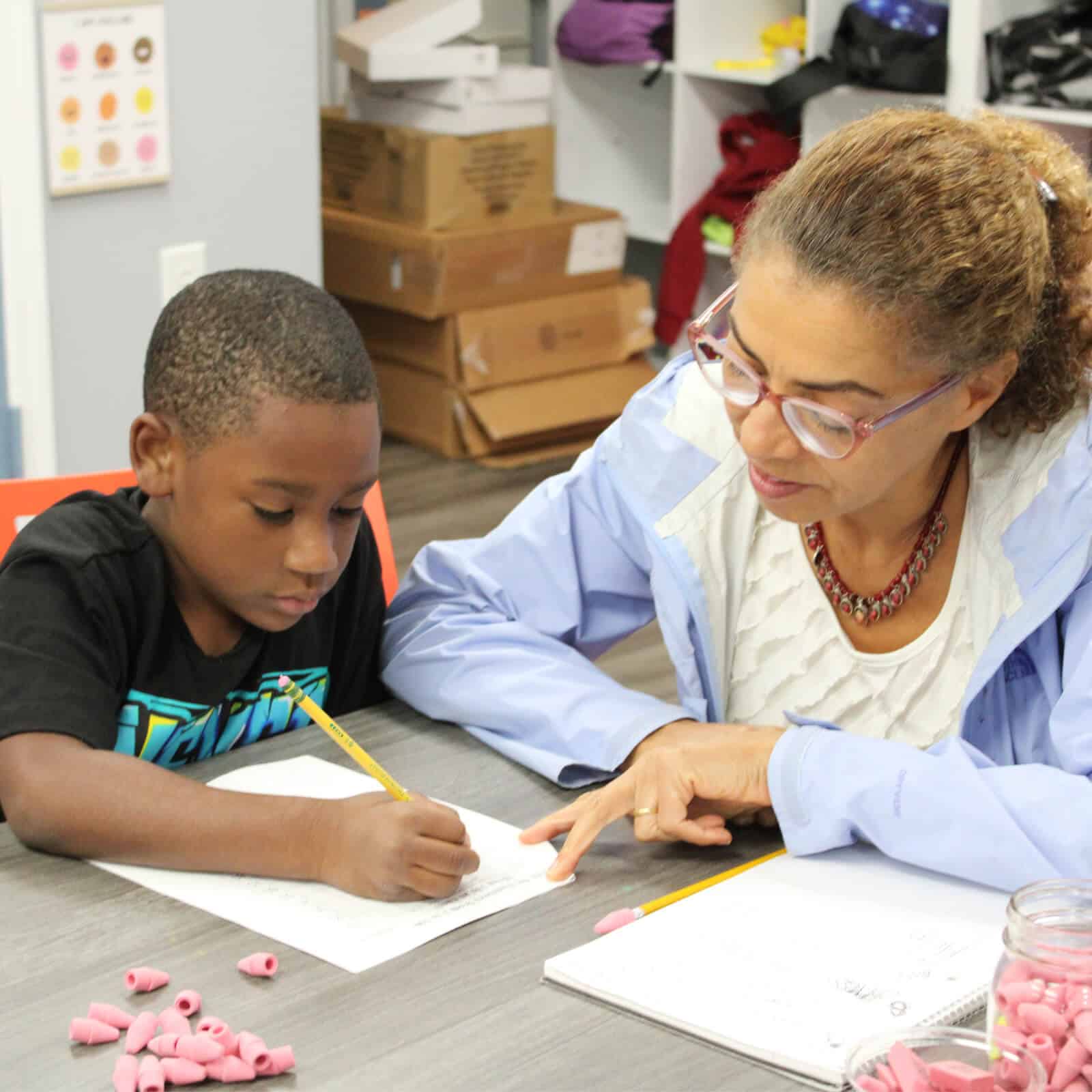 A woman sits beside a young boy, helping him with his writing assignment during a Resident Services activity. They both hold pencils, with pink beads scattered on the table and boxes and supplies visible in the background.