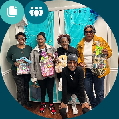 Five women, four standing and one kneeling in front, smile and hold gift baskets and bags in front of blue curtains with ribbons. It looks like a Resident Services celebration at a cheerful indoor event.
