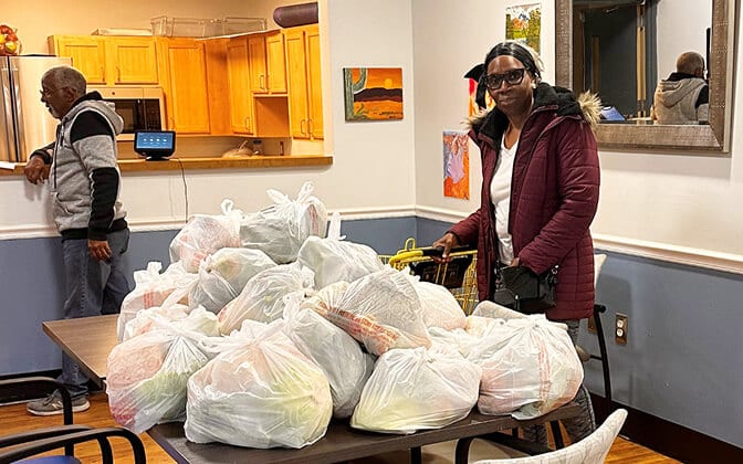 A woman in a maroon jacket stands beside a table filled with plastic bags of groceries. A man is in the background near a kitchen area. The room has colorful artwork on the walls.
