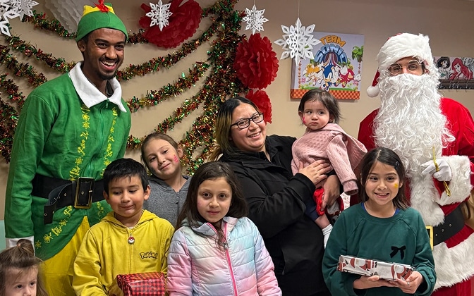 A group of smiling children and adults pose indoors with a person dressed as Santa Claus and another as an elf, surrounded by festive holiday decorations and holding wrapped presents.