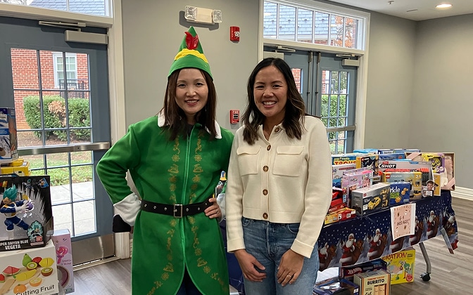Two women smiling indoors, one dressed in a green elf costume and the other in a white jacket. Behind them are tables filled with assorted toys and games. Large windows let in natural light.