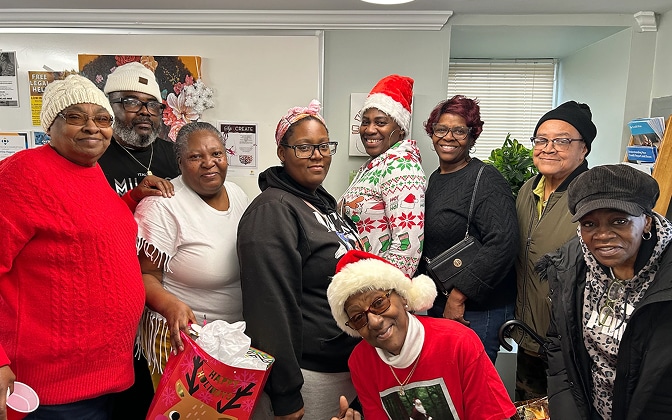A group of nine adults smile together indoors, some wearing festive holiday sweaters, Santa hats, or reindeer antlers, with one person holding a holiday gift bag.