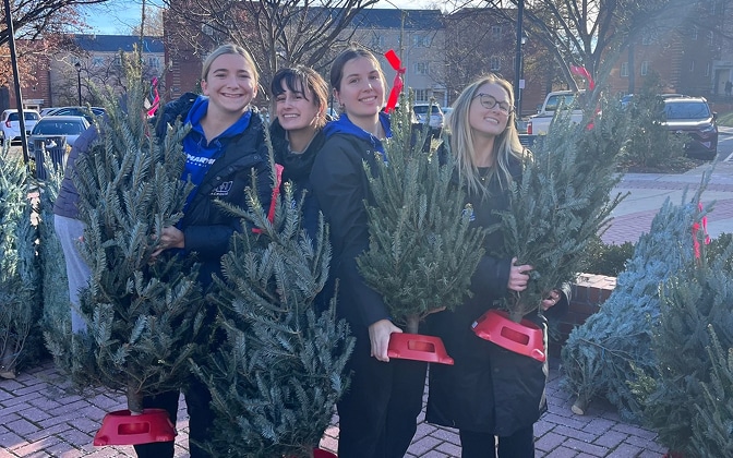 Four smiling young women stand outdoors holding small Christmas trees in red stands, with more trees around them and buildings visible in the background on a sunny day.