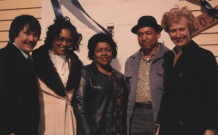 Five adults stand close together, smiling in front of a pale house door decorated with blue and white ribbon. Dressed in coats and jackets, they celebrate Affordable Homes & Communities History in a friendly and festive mood.