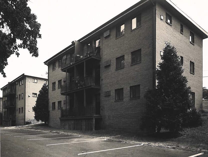 Black and white photo of a three-story brick apartment building with balconies, reflecting the history of affordable homes. Surrounded by trees, the building has empty parking spaces in front and to the side.