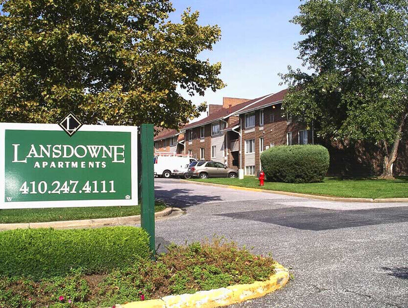 A green and white sign reading Lansdowne Apartments, 410.247.4111 stands at the entrance of this affordable homes community with brown brick buildings, cars parked, trees, and a well-kept lawn.