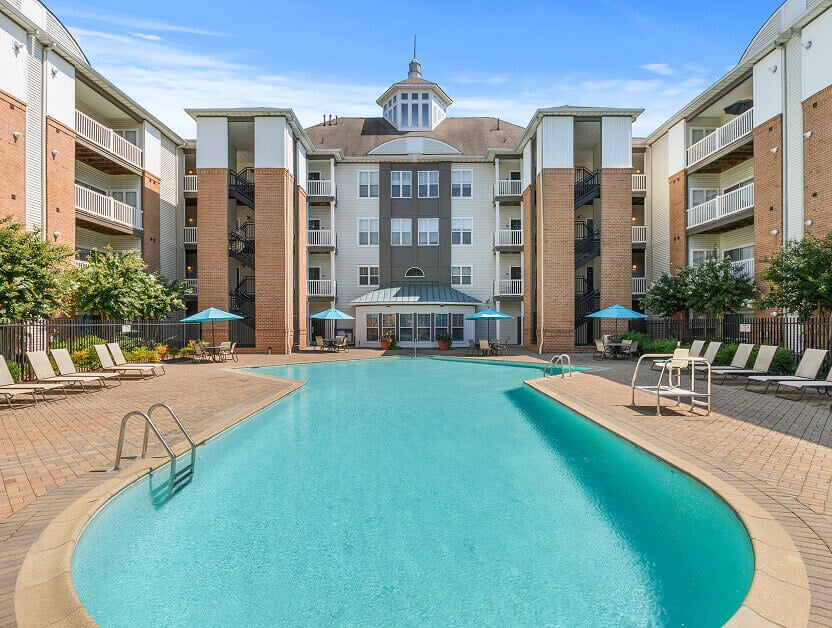Large outdoor swimming pool with lounge chairs and umbrellas, surrounded by a multi-story apartment building that reflects the rich history of affordable homes & communities, featuring balconies, brick columns, and a central pavilion under a blue sky.