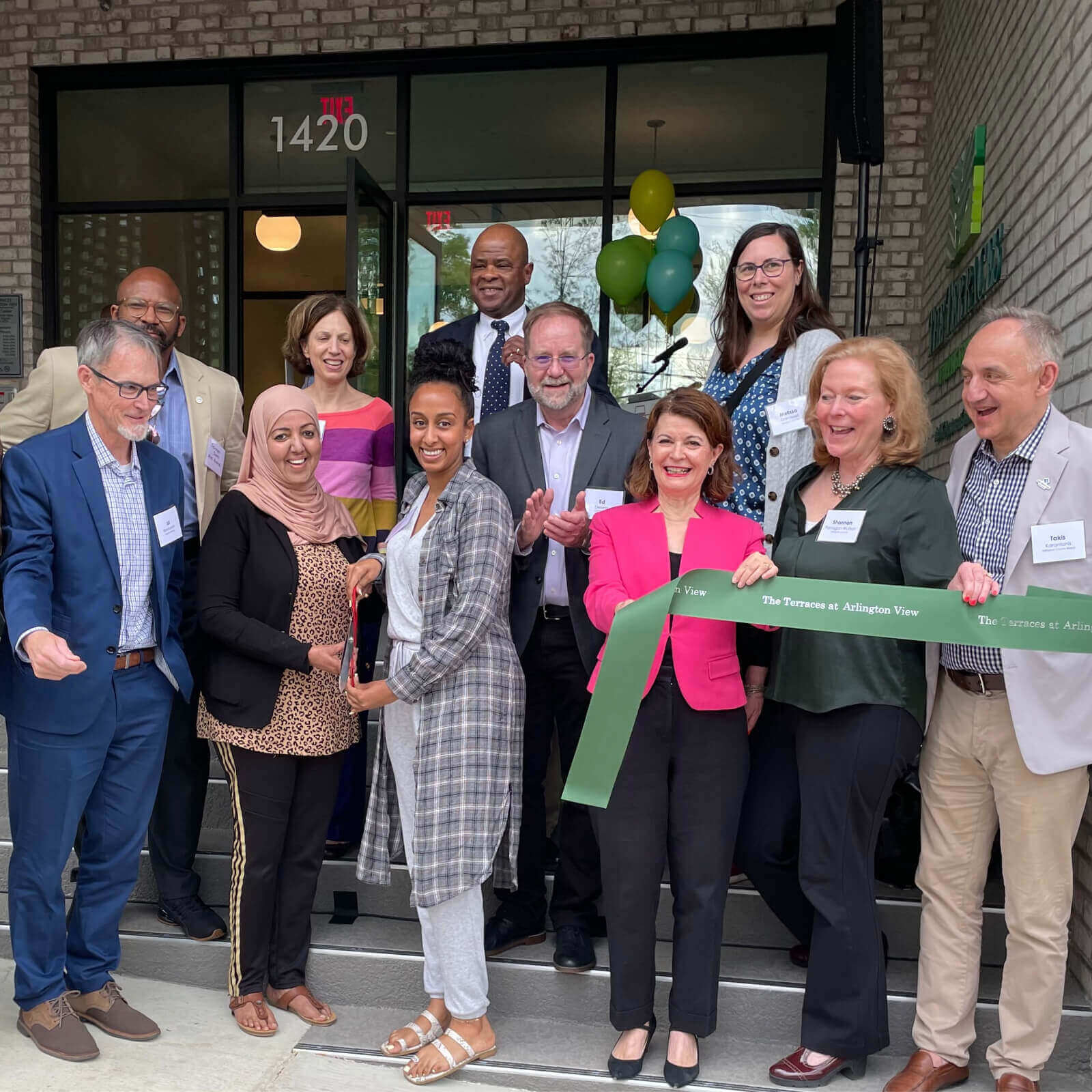 A diverse group of people smiles at a ribbon-cutting ceremony outside a building entrance, celebrating Affordable Homes & Communities History, with green and yellow balloons in the background. A woman in front holds the large green ribbon as others gather around her.