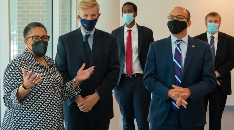 Five people wearing face masks and business attire stand indoors. One woman gestures while speaking about Building Community With AHC Welcome Baskets, while four men listen attentively in a professional or official setting.