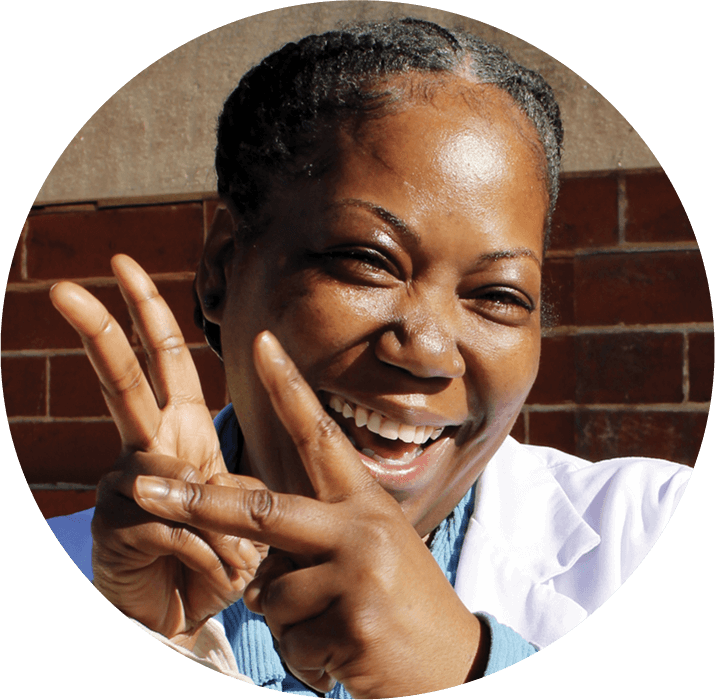 A smiling woman with braided hair, wearing a white coat, holds up her fingers in a playful peace sign gesture against a brick wall background, celebrating the achievements highlighted in the AHC Impact Report 2024.