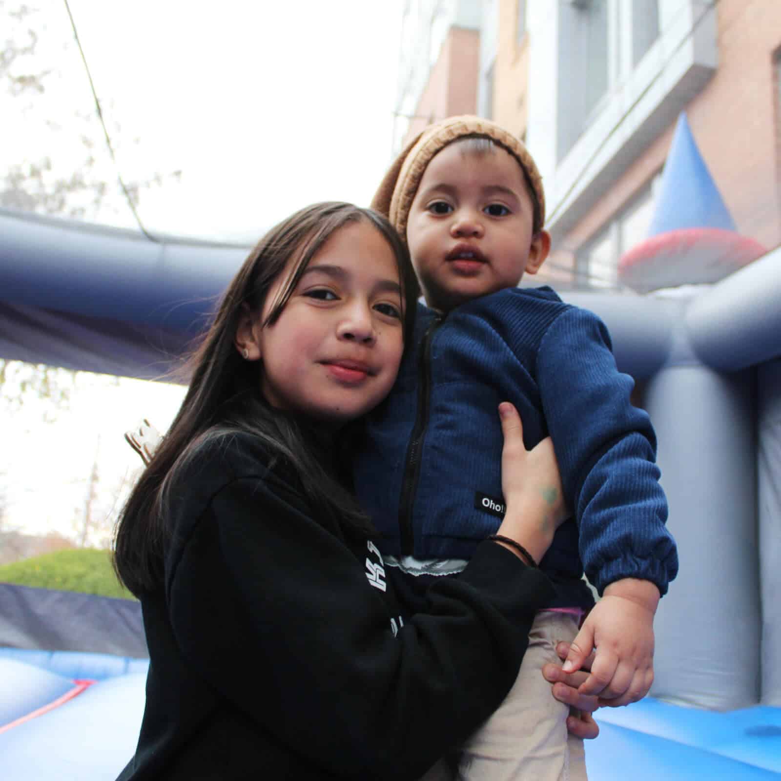 Two children pose outdoors in front of an inflatable structure. The older, in a black sweatshirt, holds the younger in a blue jacket and tan knit hat. Both smile gently at the camera—reminding us why it matters to donate to Affordable Homes & Communities.
