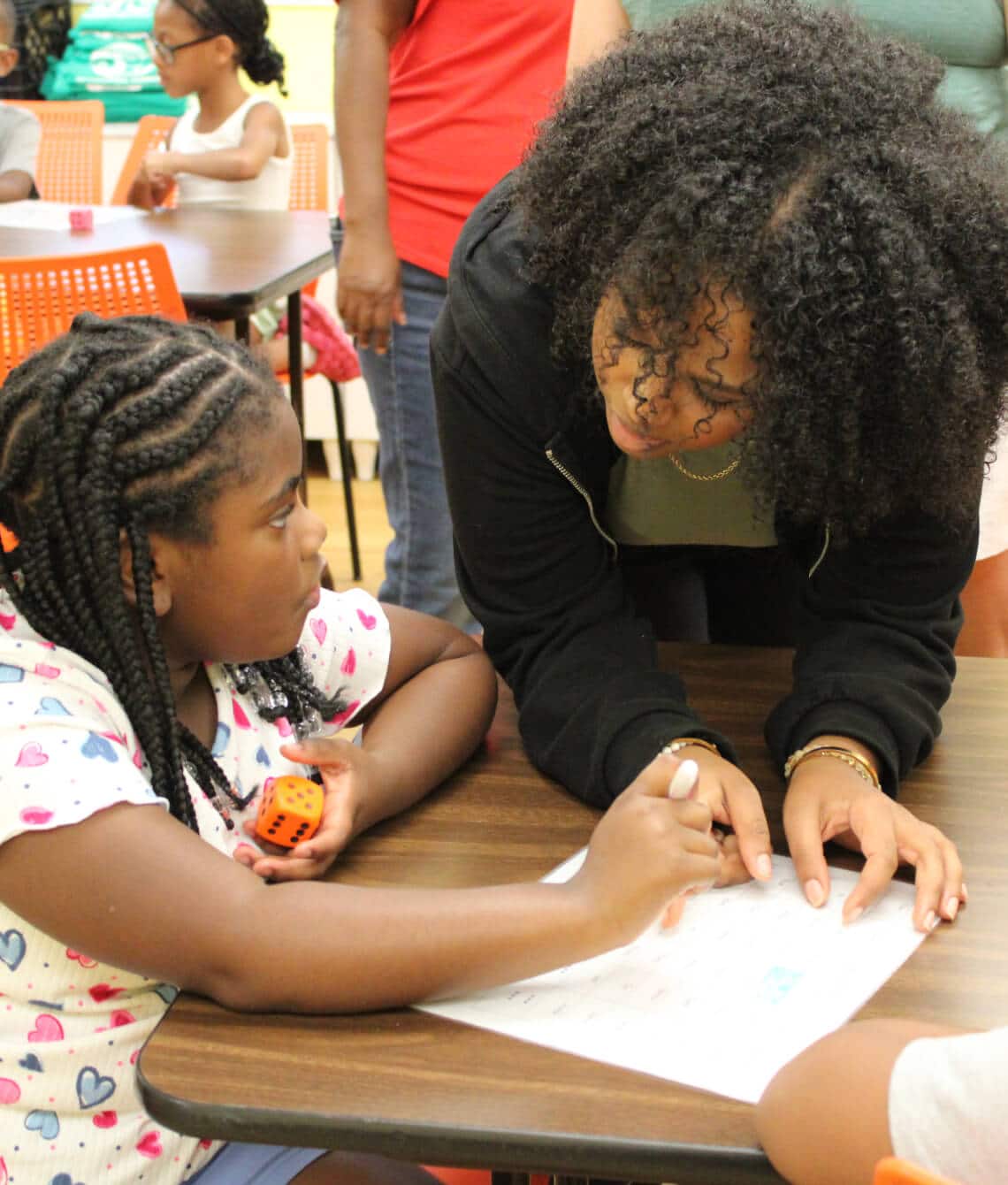 A woman leans over a table, helping a young girl with braided hair who is holding a dice and writing on a worksheet. Support moments like these when you donate to Affordable Homes & Communities. Other children and adults are in the classroom background.