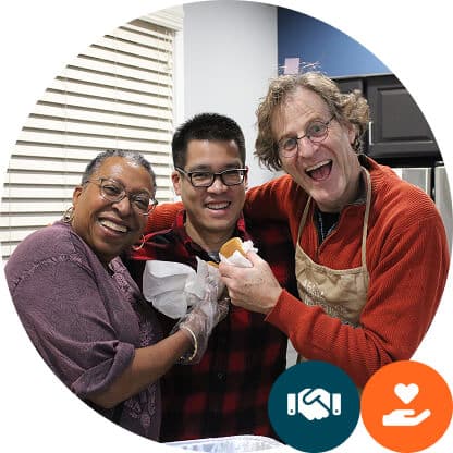 Three people smiling and standing close together in a kitchen, preparing food. One holds a sandwich, and another wears an apron. Icons of a handshake and a heart with a hand are in the corner.