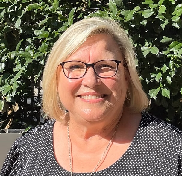 A smiling woman with blonde hair and glasses stands outdoors in front of green leafy bushes, wearing a black and white polka dot top and a necklace. Sunlight shines on her face.
