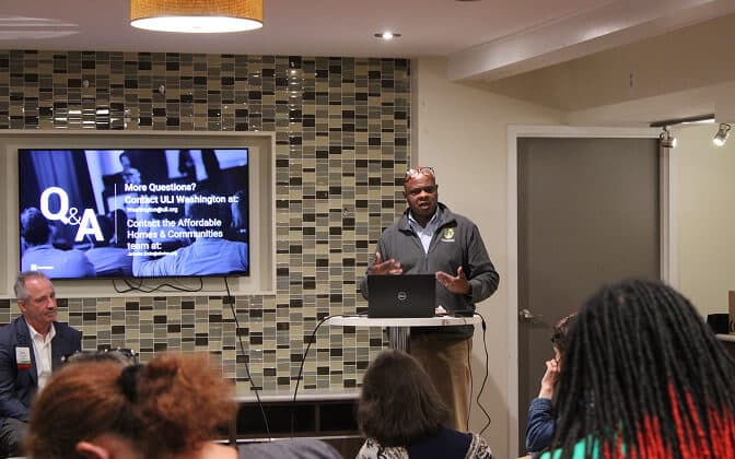 A man speaks at a podium with a laptop during a Q&A session on AHC Taps ULI for Future Planning at Serrano, while attendees listen. A screen behind him displays contact information in a modern room with patterned tile walls.