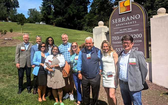 A group of eleven people stand smiling in front of the Serrano Apartments sign on a sunny day, celebrating as AHC taps ULI for future planning at Serrano, posing together on a grassy lawn with trees in the background.