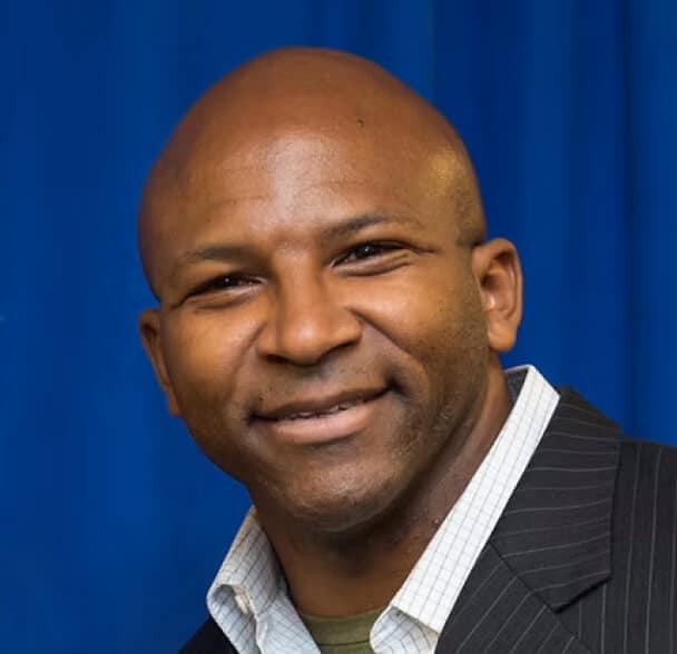 A man with a shaved head is smiling. He is wearing a white collared shirt under a dark pinstripe blazer, and the background is blue.