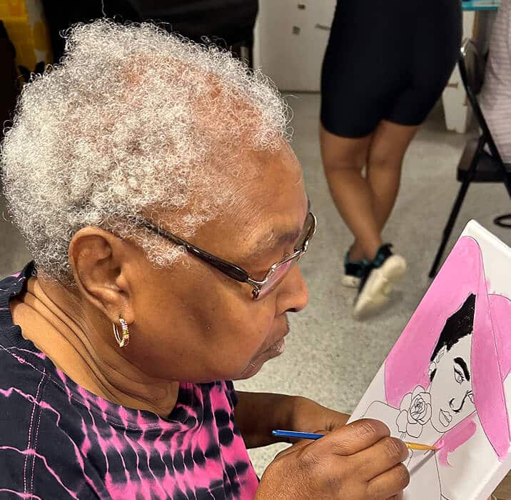 An elderly woman with short curly gray hair and glasses paints a portrait on canvas. The artwork features a womans face with a pink background. Another person stands in the background, mostly out of frame.