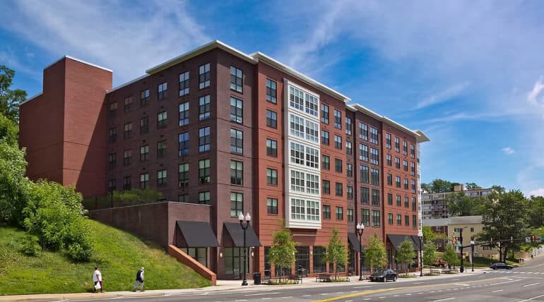 Modern mid-rise apartment building with large windows and a red brick exterior on a city street. Trees and greenery line one side, while several people—including participants from the Arlington County police tutoring program—walk along the sidewalk.