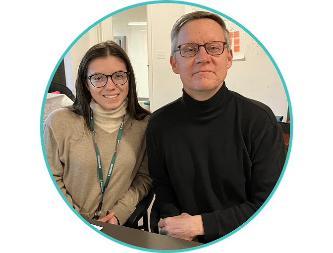 Two people wearing glasses and turtleneck sweaters sit side by side, smiling at the camera in an indoor office setting. There is a whiteboard and doorway visible in the background.