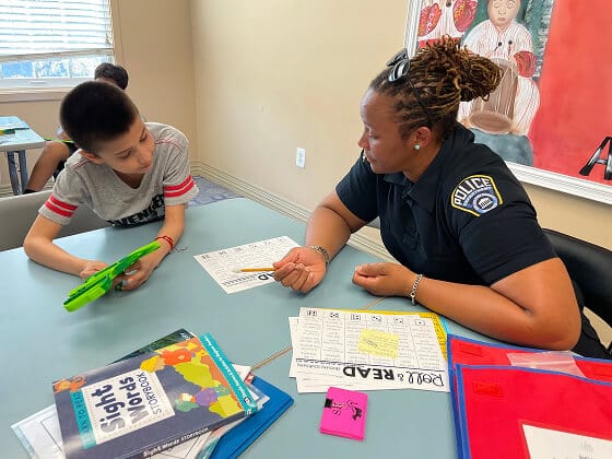 A police officer sits at a table helping a young boy with a reading worksheet in a classroom. Books and colorful folders are on the table and other students can be seen in the background.