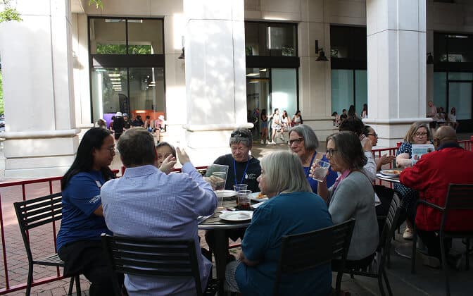 A group of people sit around a table outdoors, having a meal and talking. They are seated on black chairs, with food and drinks in front of them. The background shows a building with large white columns and windows.