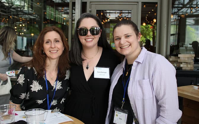 Three women smiling and posing together at a table in a bright indoor space with glass walls. One woman wears sunglasses and a name tag; all have blue lanyards. Drinks and papers are on the table in front of them.