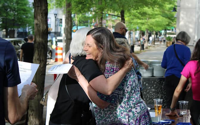 Two women smile and hug outdoors at a gathering, surrounded by other people. Green trees and city street are visible in the background, with sunlight filtering through the leaves.