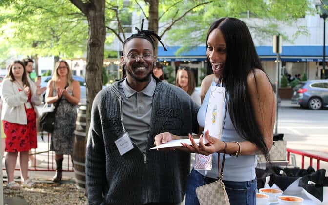 Two people stand outside smiling, one holding an open card and looking surprised. Other people in the background clap and watch. It appears to be a celebratory event with trees and parked cars nearby.
