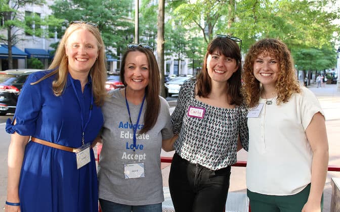 Four women smiling and posing together outdoors on a sunny day. One wears a name badge, another wears a shirt with advocacy words, and trees and parked cars are visible in the background.