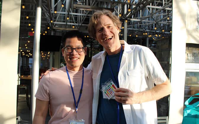 Two people stand side by side smiling outdoors, both wearing lanyards with name tags. One person is holding several colorful cards. String lights and a glass ceiling structure are visible in the background.