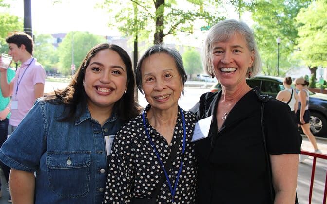 Three women smiling outdoors, standing close together. The woman on the left wears a blue shirt, the middle woman wears a polka dot blouse, and the woman on the right wears a black top. Trees and other people are in the background.