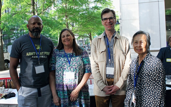 Four adults wearing name badges stand together outdoors, smiling at the camera. Trees and buildings are visible in the background, suggesting a casual or professional gathering on a sunny day.
