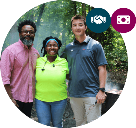 Three people stand together smiling outdoors in a wooded area. Two icons appear at the top right: a handshake (partnership) and a money symbol (funding), reflecting the impact of Affordable Homes & Communities Giving.