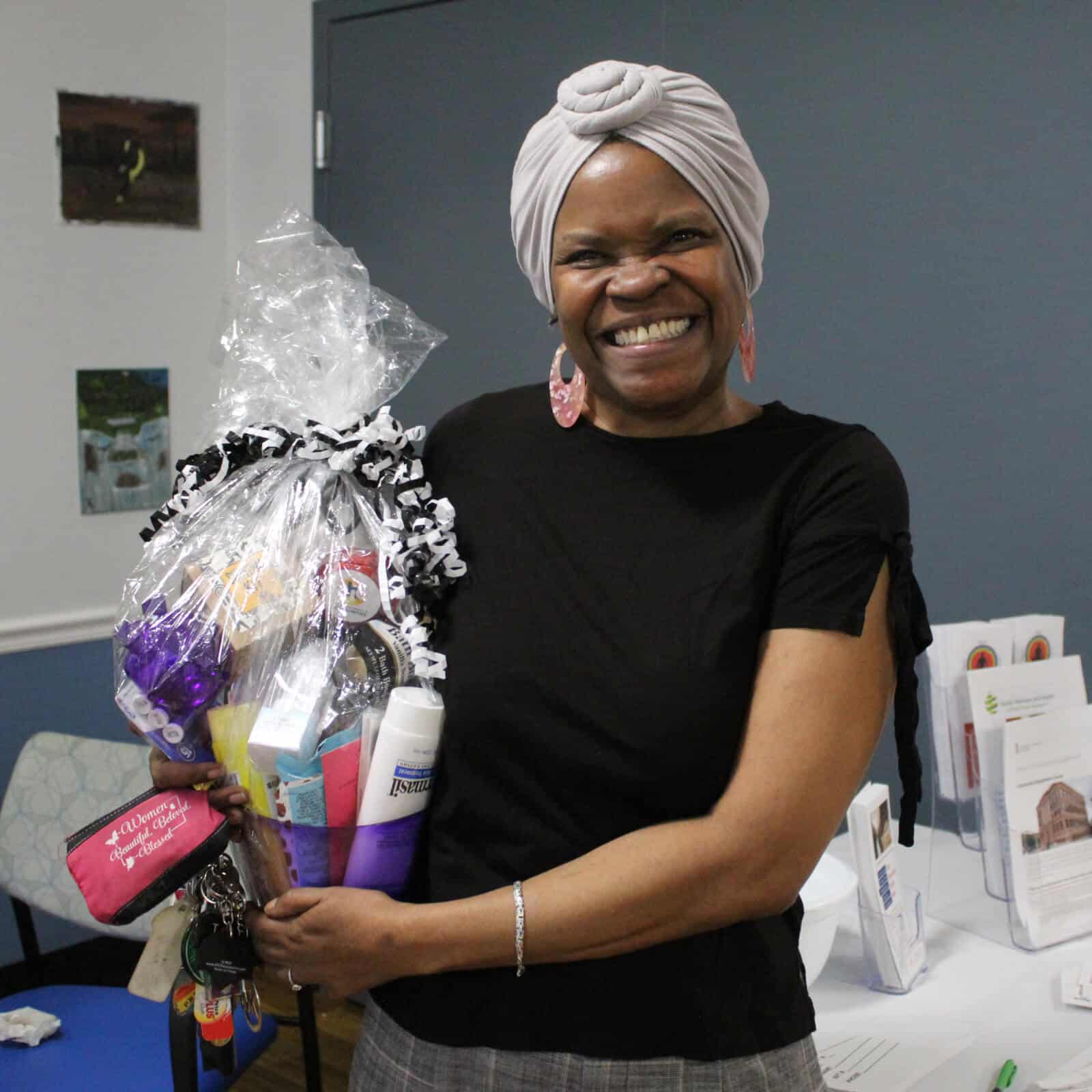 A smiling woman wearing a headwrap and black shirt holds a gift basket wrapped in cellophane. She stands indoors near a table with brochures, representing Affordable Homes & Communities Giving.