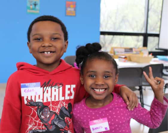 Two smiling children stand side by side, each wearing a name tag at an Affordable Homes & Communities Giving event. The boy in a red hoodie has his arm around the girl in a pink shirt, who is making a peace sign. Tables and chairs fill the background.